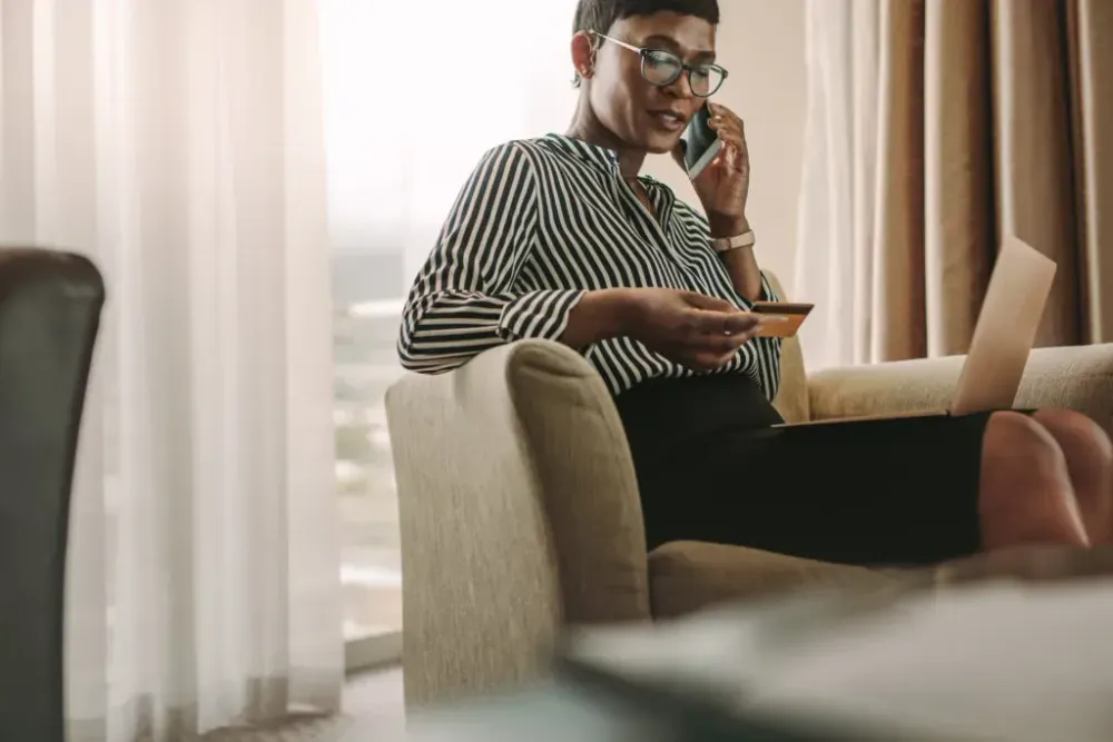 Woman in business attire sitting on the couch with a laptop in her lap, on the phone and looking at a credit card she holds in her hand
